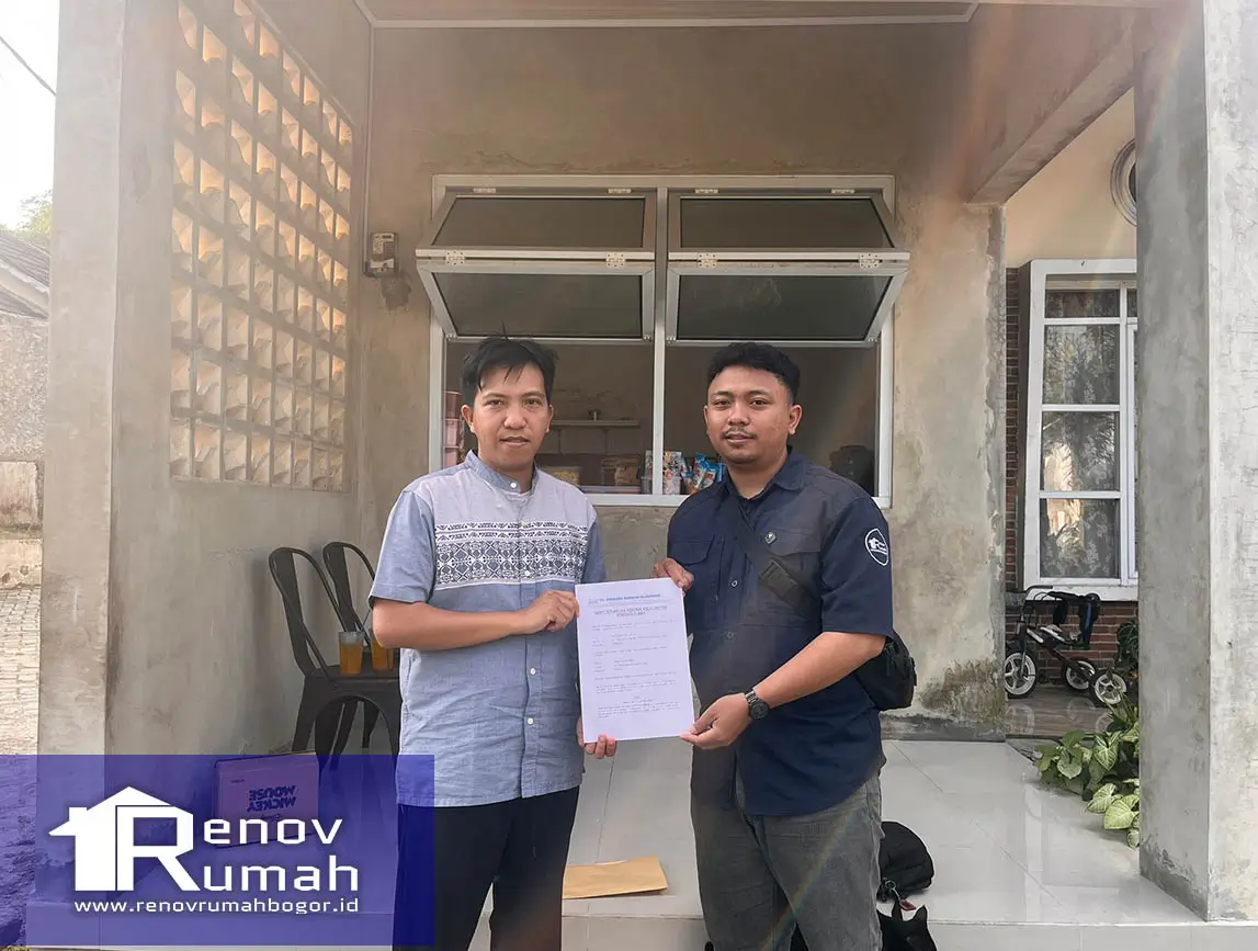 Two men stand facing the camera as one hands a document to the other in front of a house renovation setup, with a blue logo bottom-left reading 'Renov Rumah'.