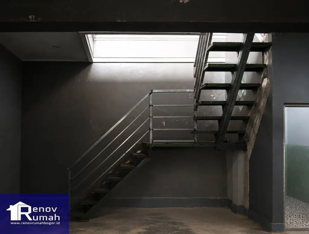 Industrial stairwell with metal staircase and railing, dark walls, and a skylight above; Renov Rumah logo bottom-left (blue panel).